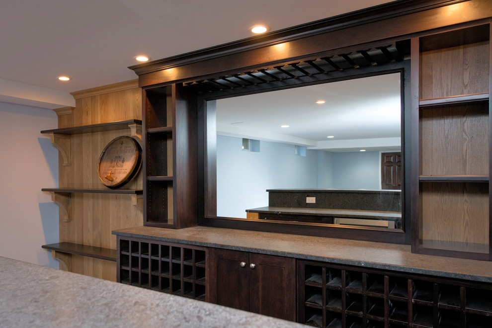 Large transitional galley seated home bar in New York with dark wood cabinets, soapstone benchtops, mirror splashback, dark hardwood floors, brown floor and black benchtop.