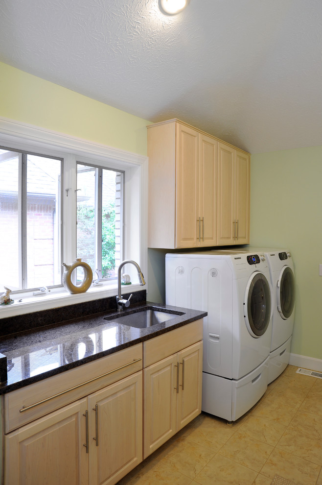 Mid-sized contemporary single-wall dedicated laundry room in Cincinnati with a single-bowl sink, raised-panel cabinets, light wood cabinets, granite benchtops, green walls, ceramic floors, a side-by-side washer and dryer and beige floor.