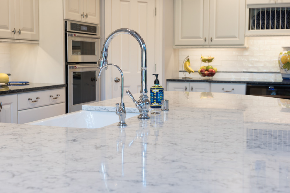 Example of a large transitional single-wall light wood floor and brown floor open concept kitchen design with a farmhouse sink, raised-panel cabinets, white cabinets, quartz countertops, white backsplash, ceramic backsplash, stainless steel appliances, an island and white countertops
