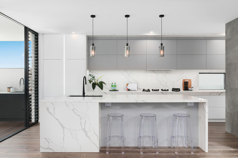 Mid-sized contemporary galley open plan kitchen in Sydney with a double-bowl sink, flat-panel cabinets, grey cabinets, marble benchtops, white splashback, marble splashback, black appliances, light hardwood floors, multiple islands, white benchtop and brown floor.