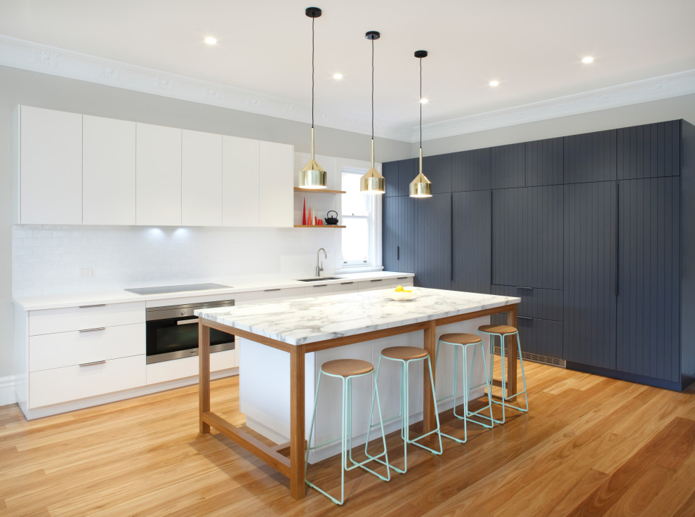 Inspiration for a large contemporary l-shaped open plan kitchen in Sydney with a double-bowl sink, flat-panel cabinets, white cabinets, white splashback, panelled appliances, medium hardwood floors, with island, beige floor and white benchtop.