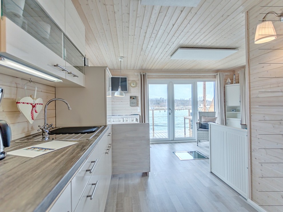 This is an example of a beach style single-wall open plan kitchen in Hamburg with a drop-in sink, flat-panel cabinets, white cabinets, white splashback, timber splashback, stainless steel appliances, laminate floors, no island and grey floor.