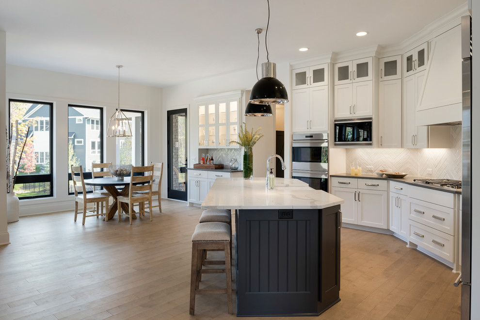 Example of a transitional l-shaped light wood floor and beige floor eat-in kitchen design in Minneapolis with a farmhouse sink, shaker cabinets, white cabinets, white backsplash, stainless steel appliances, an island and gray countertops