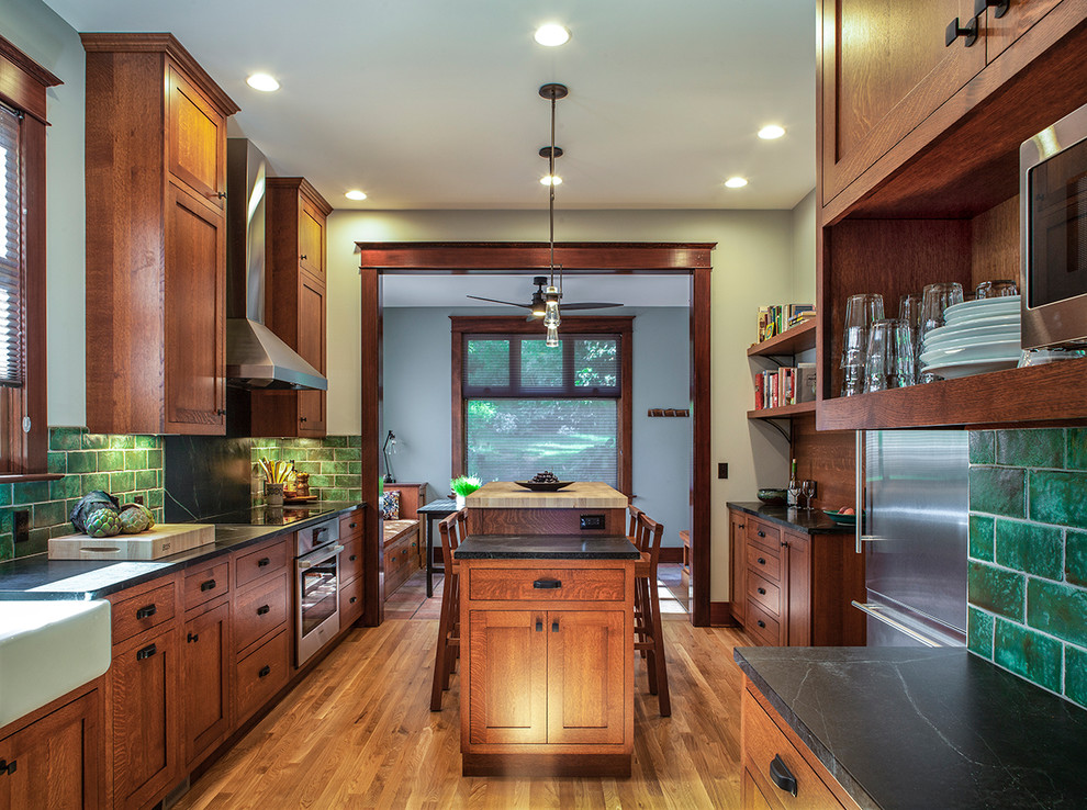 Large arts and crafts galley separate kitchen in Portland with a farmhouse sink, shaker cabinets, soapstone benchtops, green splashback, ceramic splashback, stainless steel appliances, medium hardwood floors, with island, medium wood cabinets, brown floor and black benchtop.