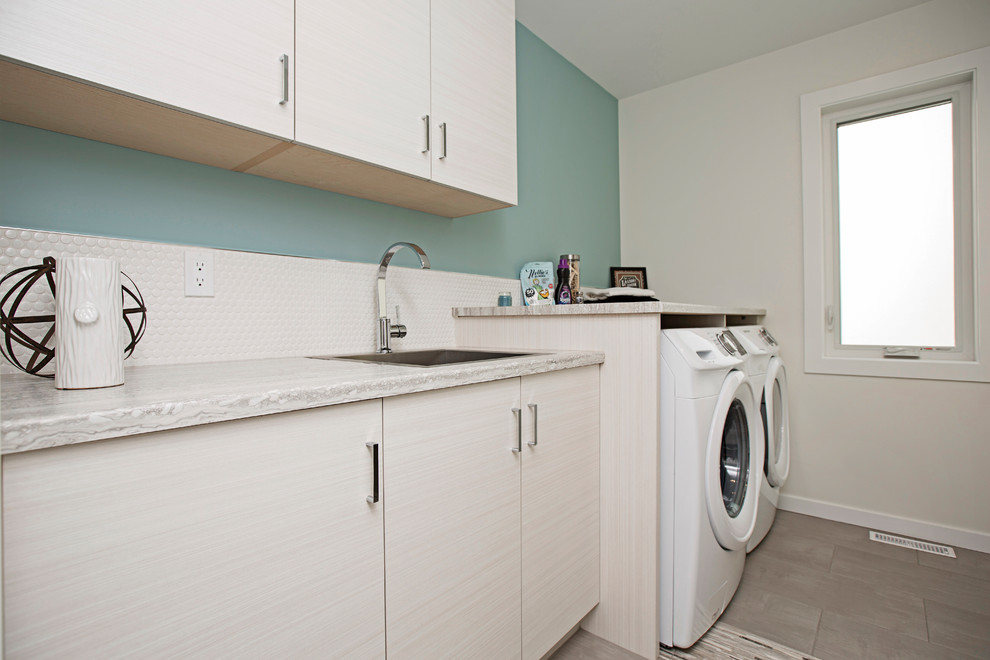 This is an example of a mid-sized contemporary single-wall dedicated laundry room in Edmonton with a drop-in sink, flat-panel cabinets, light wood cabinets, laminate benchtops, blue walls, ceramic floors, a side-by-side washer and dryer and beige floor.