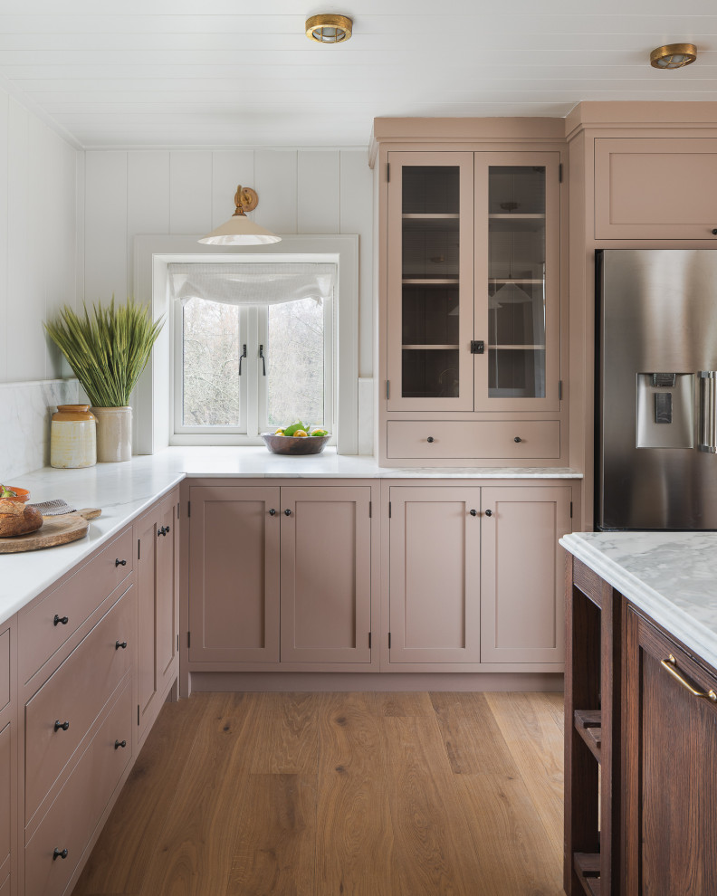 This is an example of a mid-sized country kitchen in Other with with island, a farmhouse sink, recessed-panel cabinets, pink cabinets, granite benchtops, white splashback, granite splashback, stainless steel appliances, light hardwood floors and white benchtop.