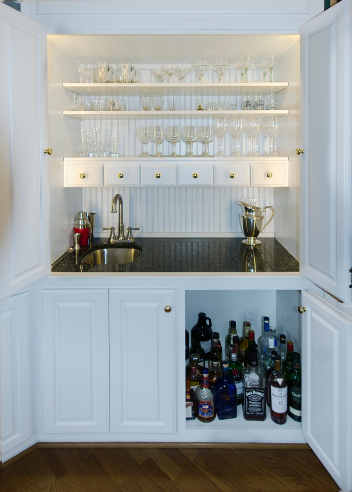 Photo of a small traditional wet bar in DC Metro with an undermount sink, raised-panel cabinets, white cabinets, granite benchtops and medium hardwood floors.