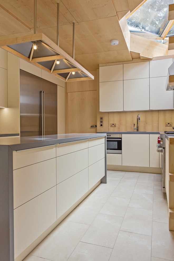 This is an example of a mid-sized contemporary u-shaped eat-in kitchen in Hertfordshire with flat-panel cabinets, solid surface benchtops, timber splashback, stainless steel appliances, limestone floors, with island, white floor and beige cabinets.