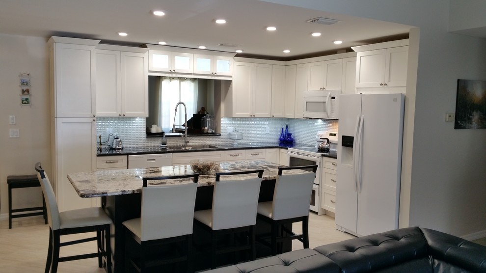 Photo of a mid-sized transitional l-shaped open plan kitchen in Tampa with an undermount sink, shaker cabinets, white cabinets, granite benchtops, white splashback, ceramic splashback, white appliances, porcelain floors and with island.