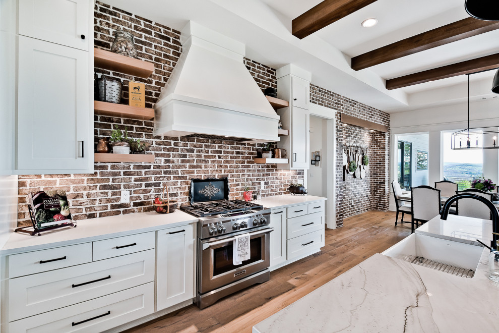 Large farmhouse u-shaped kitchen/diner in Other with a belfast sink, raised-panel cabinets, white cabinets, engineered stone countertops, red splashback, brick splashback, stainless steel appliances, light hardwood flooring, an island, white worktops and exposed beams.