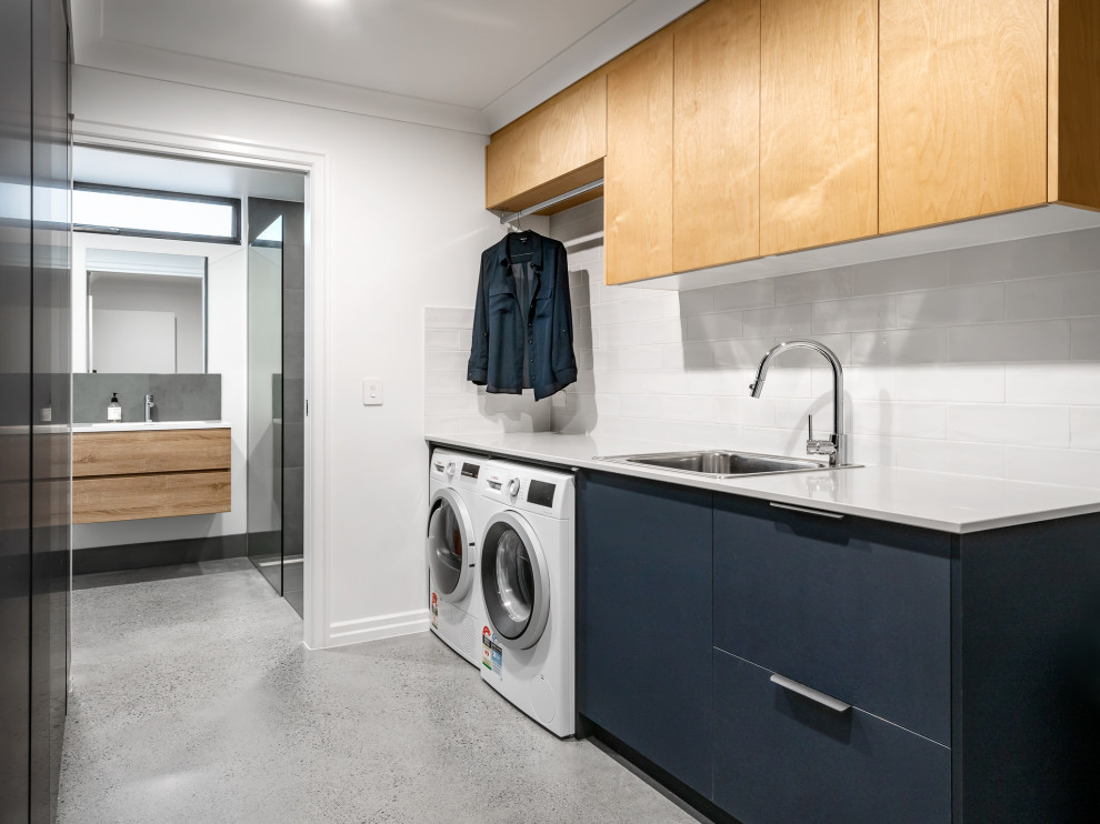 Foto på en funkis vita tvättstuga, med en undermonterad diskho, släta luckor, blå skåp, vitt stänkskydd, vita väggar, betonggolv, en tvättmaskin och torktumlare bredvid varandra och grått golv