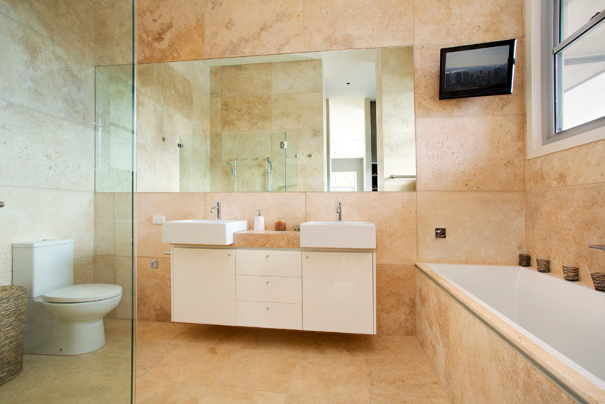 Photo of a large country master bathroom in Sydney with a drop-in tub, a double shower, a two-piece toilet, stone tile, travertine floors, flat-panel cabinets, white cabinets, beige tile, beige walls, a trough sink, solid surface benchtops, beige floor, a hinged shower door and beige benchtops.
