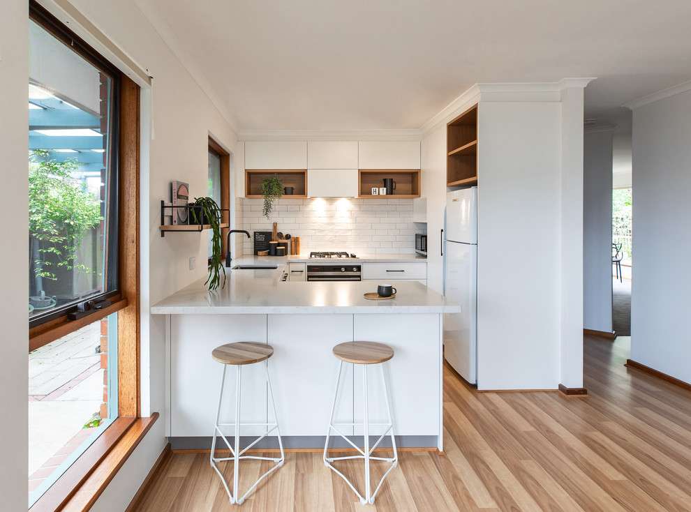 This is an example of a scandinavian u-shaped kitchen in Adelaide with an undermount sink, flat-panel cabinets, white cabinets, white splashback, subway tile splashback, white appliances, brown floor, white benchtop, quartz benchtops and laminate floors.