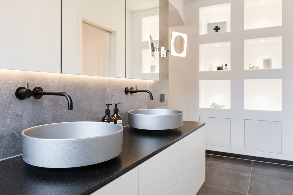 This is an example of a mid-sized contemporary bathroom in Essen with flat-panel cabinets, white cabinets, beige tile, white walls, cement tiles, a vessel sink, brown floor and black benchtops.