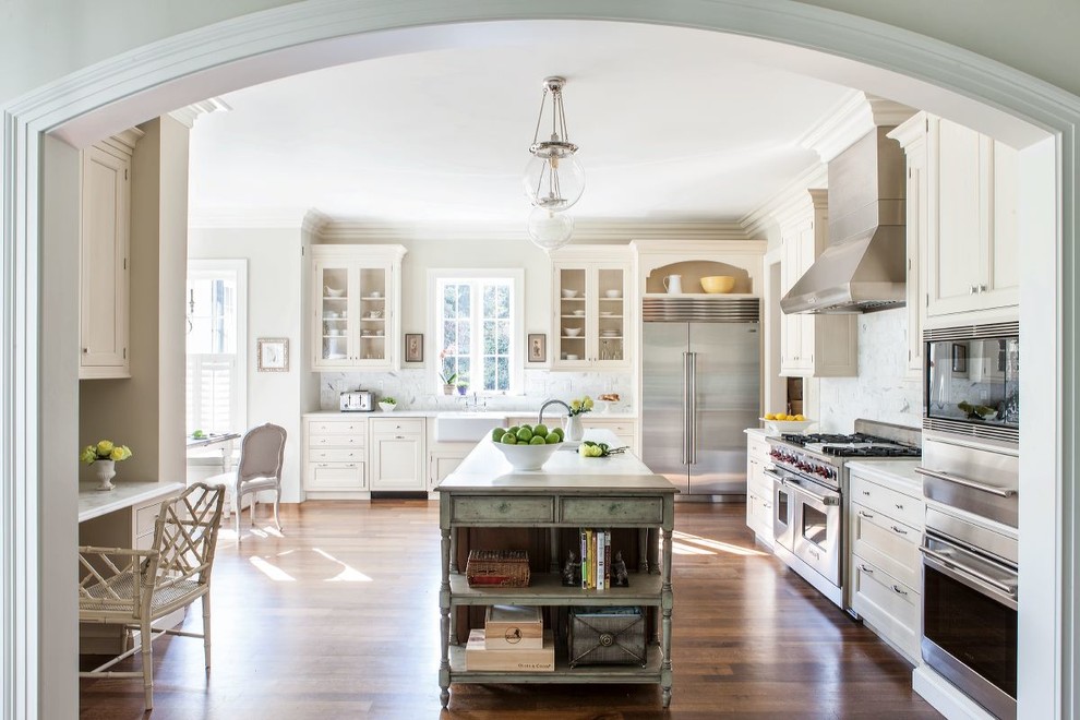 This is an example of a traditional l-shaped kitchen in DC Metro with a farmhouse sink, glass-front cabinets, white cabinets, white splashback, stone tile splashback, stainless steel appliances, medium hardwood floors and with island.