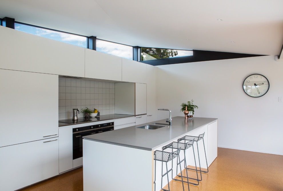 This is an example of a midcentury galley kitchen in Auckland with an undermount sink, flat-panel cabinets, white cabinets, white splashback, ceramic splashback, panelled appliances, with island, orange floor and grey benchtop.