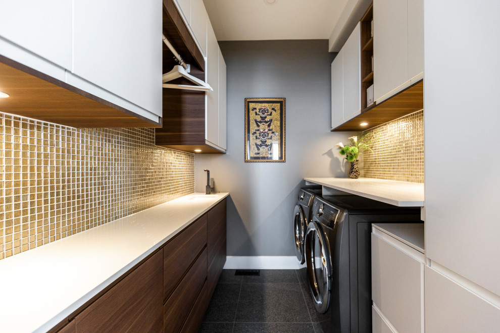 This is an example of a large contemporary galley dedicated laundry room in Montreal with an undermount sink, flat-panel cabinets, medium wood cabinets, quartz benchtops, beige splashback, glass tile splashback, grey walls, porcelain floors, a side-by-side washer and dryer, grey floor and white benchtop.