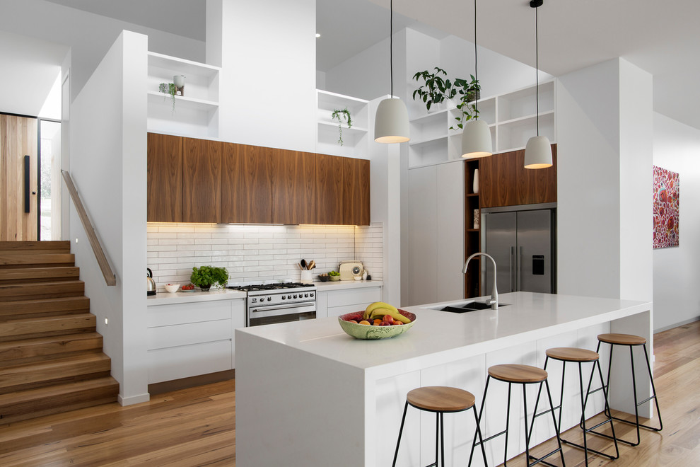 Design ideas for a contemporary l-shaped open plan kitchen in Melbourne with a double-bowl sink, flat-panel cabinets, white cabinets, white splashback, subway tile splashback, stainless steel appliances, light hardwood floors, with island and white benchtop.