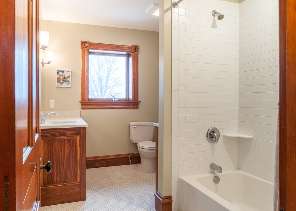 Photo of a mid-sized country master bathroom in Minneapolis with raised-panel cabinets, medium wood cabinets, an alcove tub, a shower/bathtub combo, a two-piece toilet, white tile, ceramic tile, green walls, mosaic tile floors, an undermount sink and engineered quartz benchtops.