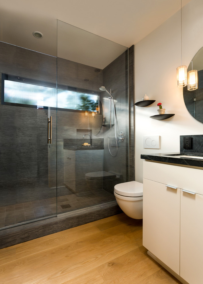 This is an example of a small midcentury 3/4 bathroom in Los Angeles with flat-panel cabinets, white cabinets, an alcove shower, a wall-mount toilet, gray tile, stone tile, white walls, light hardwood floors, an undermount sink, soapstone benchtops, yellow floor and a hinged shower door.