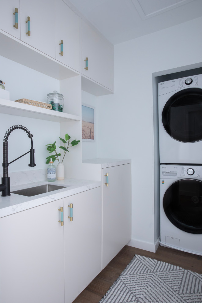 Photo of a small beach style l-shaped dedicated laundry room in Los Angeles with an undermount sink, flat-panel cabinets, white cabinets, quartz benchtops, white splashback, engineered quartz splashback, white walls, dark hardwood floors, a stacked washer and dryer and white benchtop.