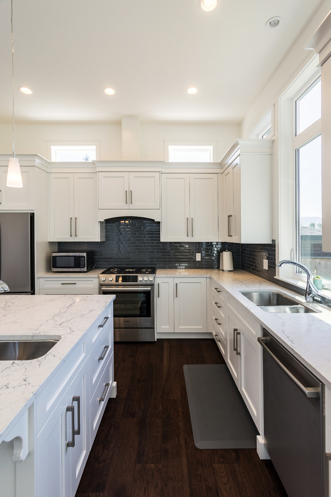 Photo of a large modern l-shaped open plan kitchen in Vancouver with a double-bowl sink, shaker cabinets, white cabinets, quartzite benchtops, black splashback, glass tile splashback, stainless steel appliances, dark hardwood floors, with island, brown floor and white benchtop.