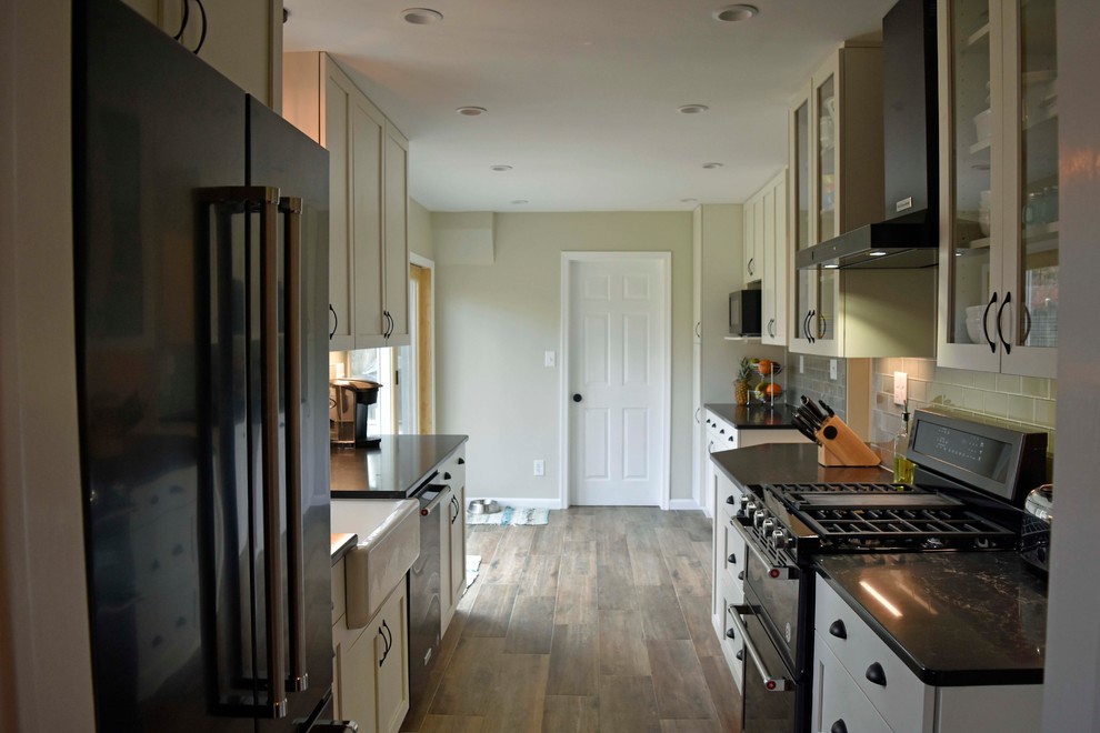 Example of a mid-sized transitional galley laminate floor and brown floor enclosed kitchen design in DC Metro with a farmhouse sink, shaker cabinets, white cabinets, granite countertops, gray backsplash, subway tile backsplash, stainless steel appliances, no island and black countertops