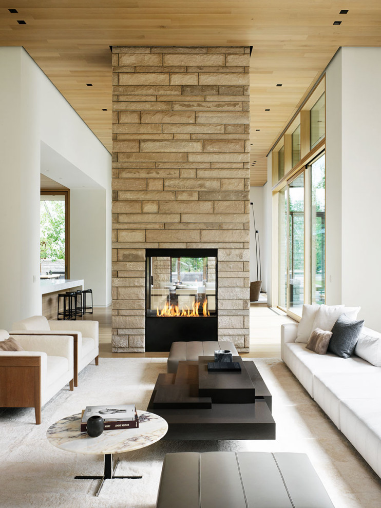 На фото: открытая гостиная комната в стиле модернизм с белыми стенами, двусторонним камином, фасадом камина из камня и деревянным потолком