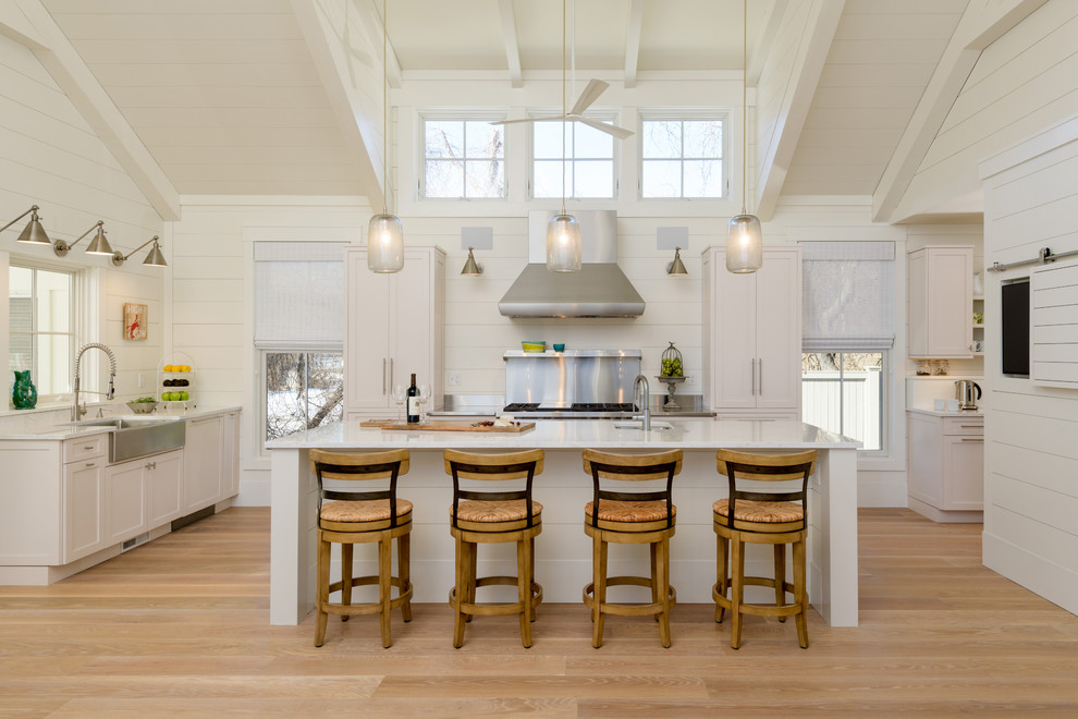 Inspiration for a beach style l-shaped kitchen in Boston with a farmhouse sink, shaker cabinets, white cabinets, white splashback, light hardwood floors, with island and beige floor.