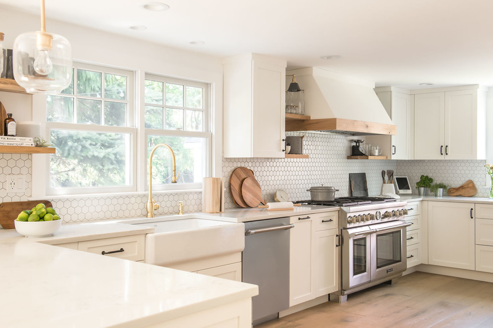 Large country u-shaped eat-in kitchen in Boston with a farmhouse sink, shaker cabinets, white cabinets, quartz benchtops, white splashback, mosaic tile splashback, stainless steel appliances, light hardwood floors, a peninsula, brown floor and white benchtop.