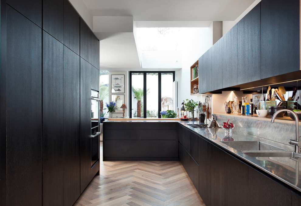 This is an example of a mid-sized contemporary l-shaped open plan kitchen in Dublin with an integrated sink, flat-panel cabinets, dark wood cabinets, stainless steel benchtops, metallic splashback, glass sheet splashback, panelled appliances, medium hardwood floors, a peninsula and multi-coloured floor.