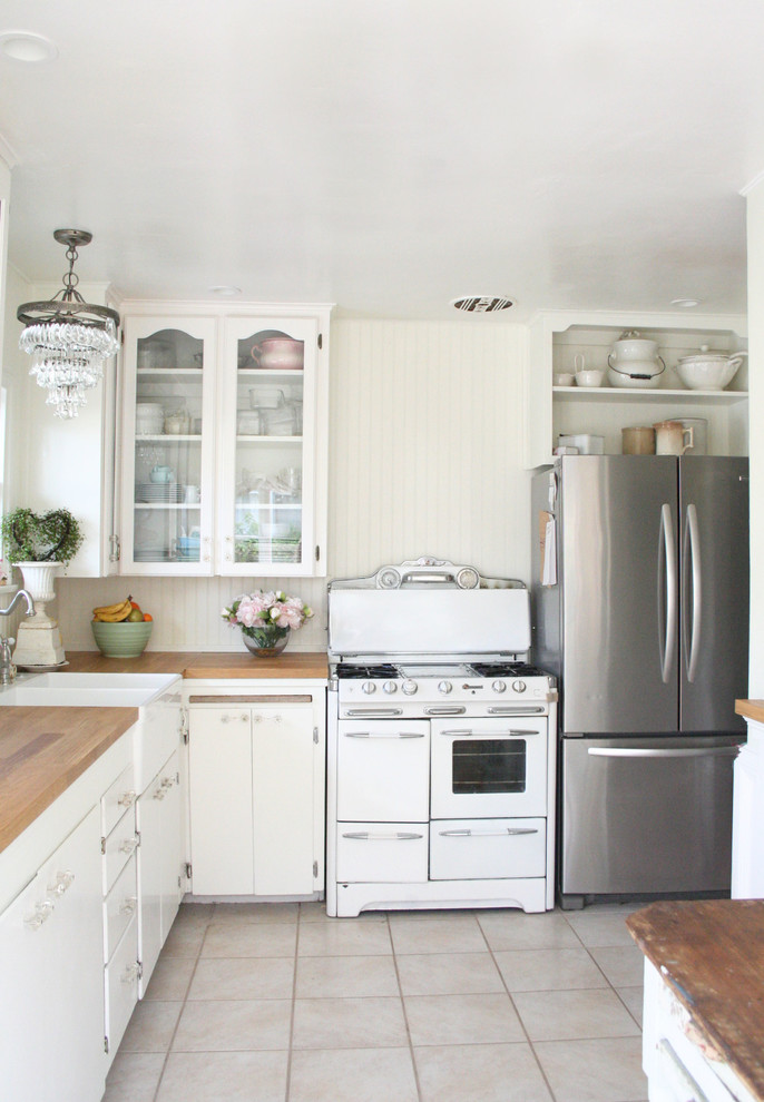 Small traditional l-shaped eat-in kitchen in Santa Barbara with a farmhouse sink, glass-front cabinets, white cabinets, wood benchtops, white splashback, stainless steel appliances, ceramic floors and no island.