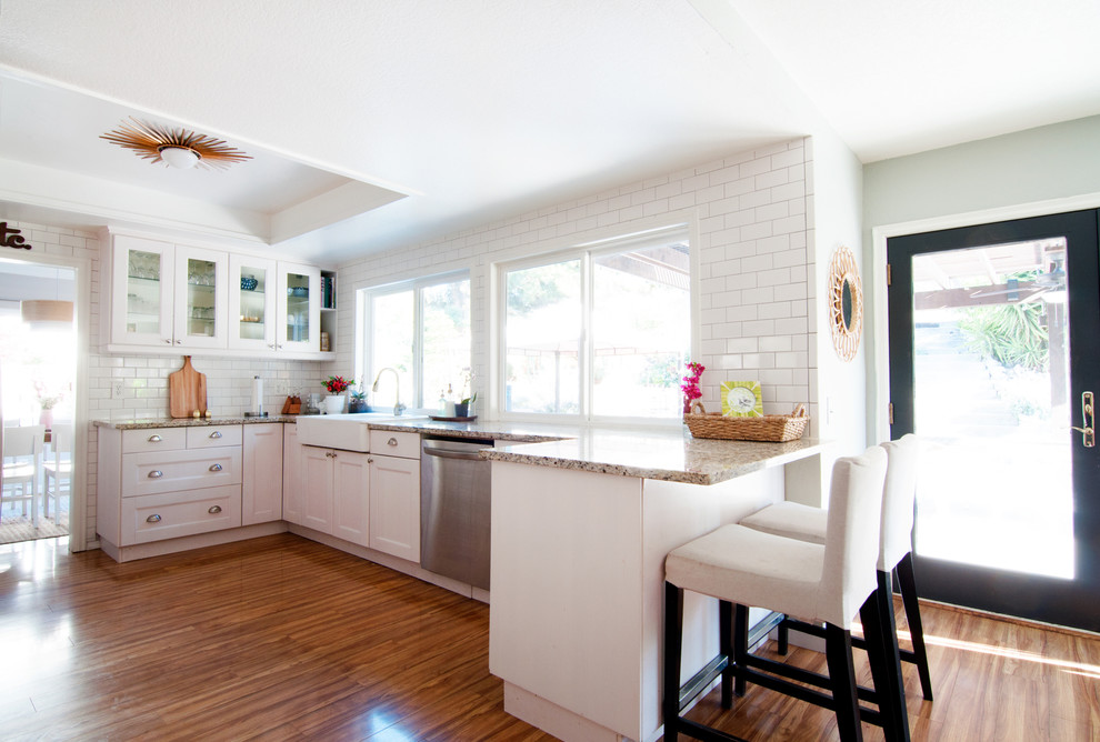 Mid-sized eclectic galley open plan kitchen in Los Angeles with a farmhouse sink, glass-front cabinets, white cabinets, granite benchtops, white splashback, subway tile splashback, stainless steel appliances and a peninsula.