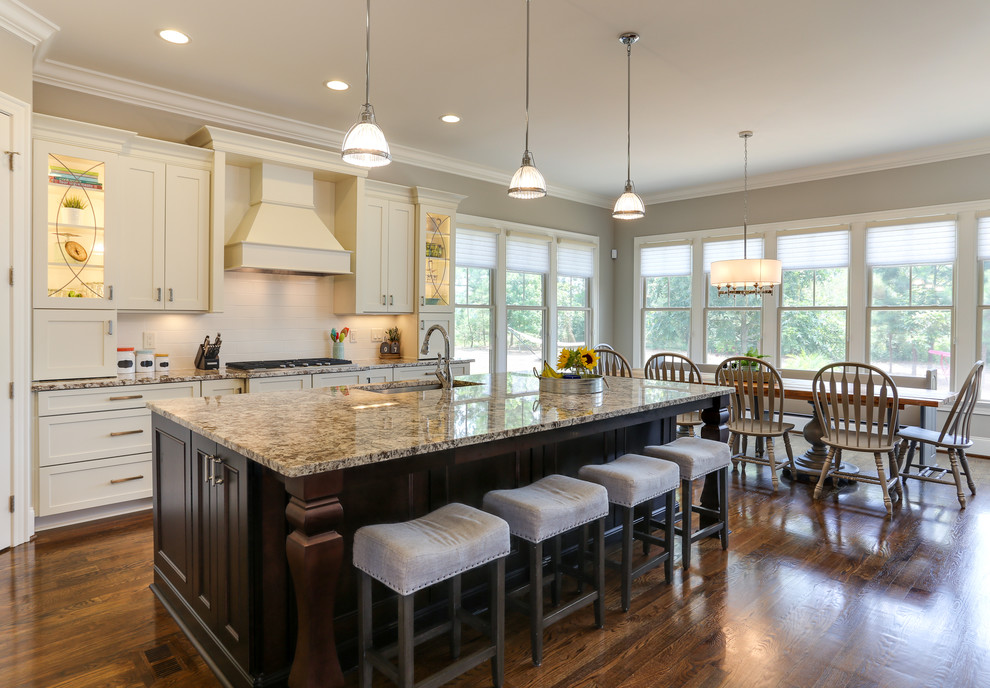 Photo of a mid-sized transitional l-shaped open plan kitchen in Raleigh with an undermount sink, recessed-panel cabinets, white cabinets, granite benchtops, white splashback, ceramic splashback, stainless steel appliances, with island, dark hardwood floors and brown floor.