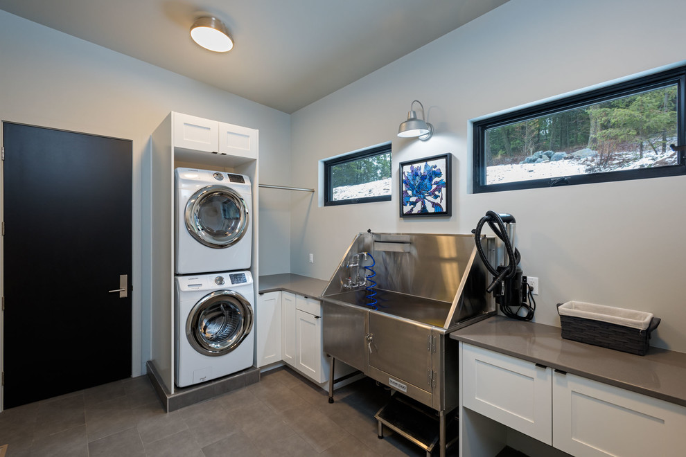 Inspiration for a mid-sized modern single-wall utility room in Seattle with recessed-panel cabinets, white cabinets, solid surface benchtops, grey walls, porcelain floors, a stacked washer and dryer and grey floor.