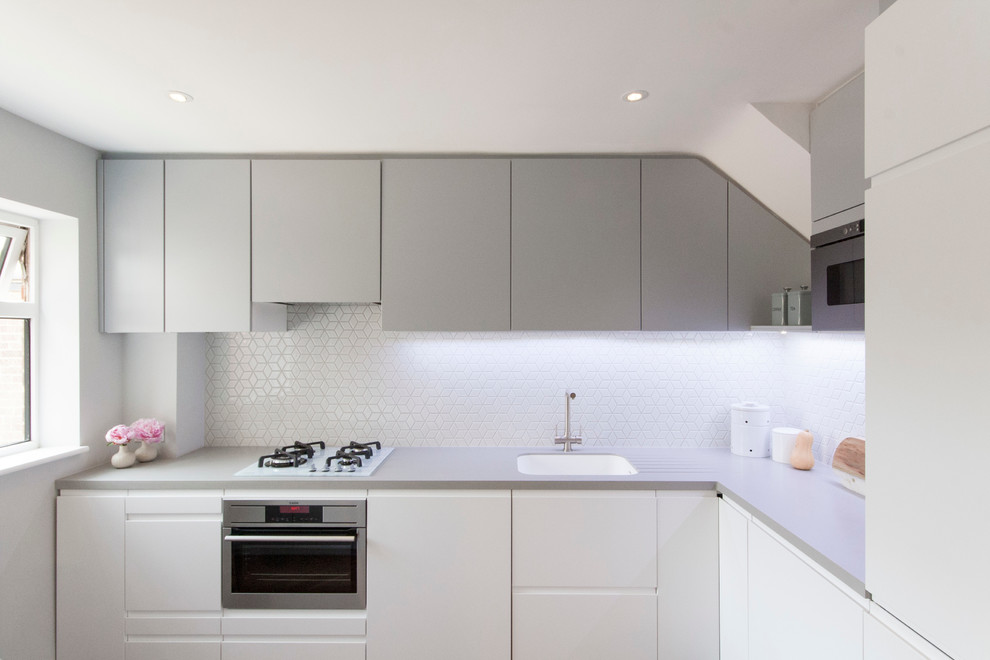 This is an example of a small contemporary l-shaped open plan kitchen in London with a drop-in sink, flat-panel cabinets, white cabinets, solid surface benchtops, white splashback, mosaic tile splashback, stainless steel appliances, dark hardwood floors and with island.