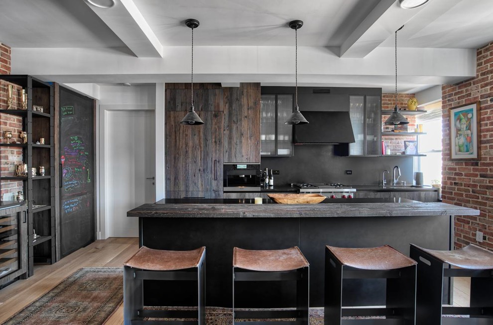 This is an example of a mid-sized industrial kitchen in New York with glass-front cabinets, black splashback, stainless steel appliances, light hardwood floors, with island and black benchtop.