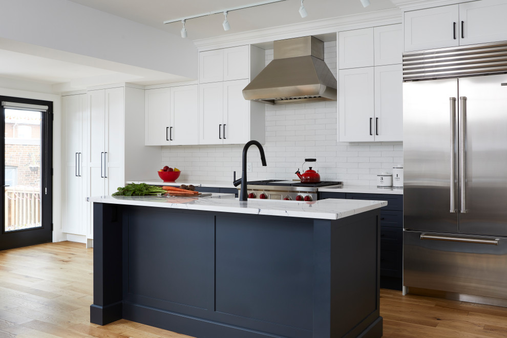 This is an example of a mid-sized transitional galley eat-in kitchen in Toronto with a farmhouse sink, shaker cabinets, white cabinets, quartz benchtops, white splashback, subway tile splashback, stainless steel appliances, light hardwood floors, with island, brown floor, white benchtop and vaulted.