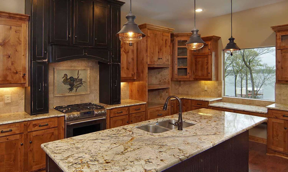 This is an example of a mid-sized arts and crafts l-shaped eat-in kitchen in Dallas with an undermount sink, recessed-panel cabinets, brown cabinets, granite benchtops, beige splashback, limestone splashback, stainless steel appliances, medium hardwood floors, with island and brown floor.