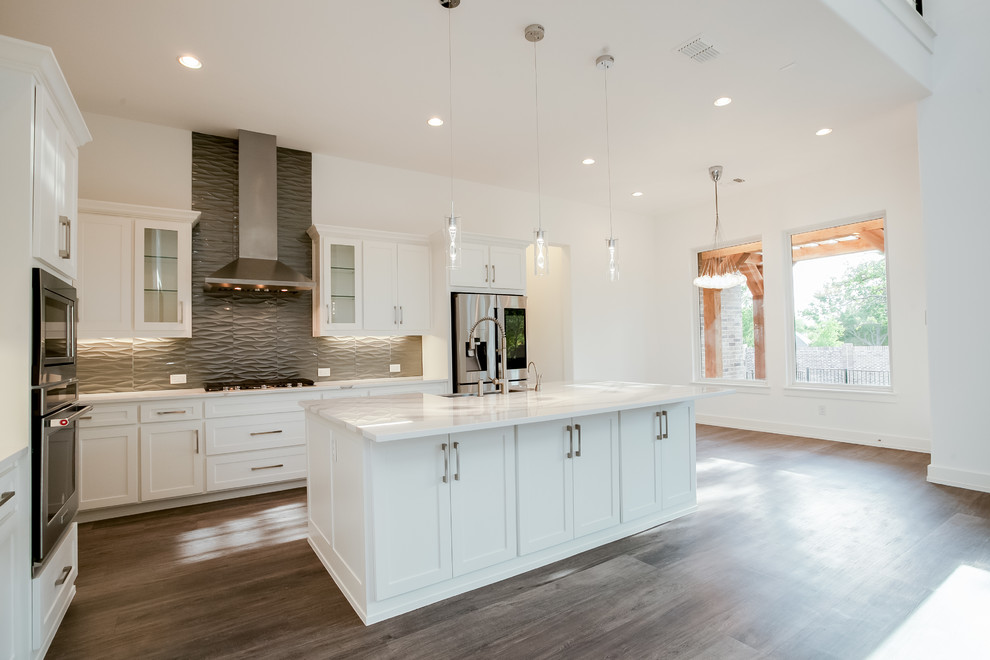 Large modern l-shaped open plan kitchen in Dallas with a farmhouse sink, recessed-panel cabinets, white cabinets, granite benchtops, grey splashback, glass tile splashback, stainless steel appliances, medium hardwood floors, with island, brown floor and white benchtop.