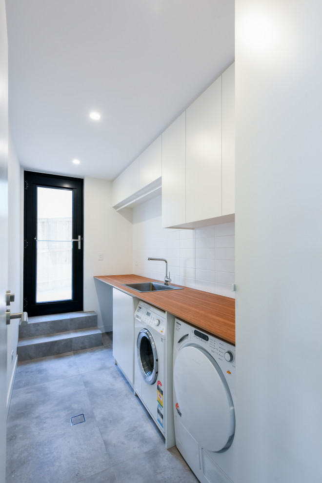 Design ideas for a large modern single-wall dedicated laundry room in Sydney with a drop-in sink, flat-panel cabinets, white cabinets, wood benchtops, white walls, ceramic floors, a side-by-side washer and dryer, grey floor and brown benchtop.