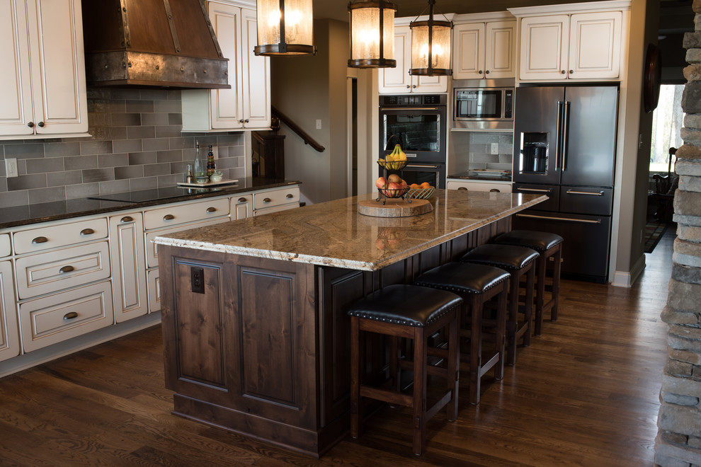 Example of a large mountain style l-shaped dark wood floor and brown floor eat-in kitchen design with raised-panel cabinets, dark wood cabinets, granite countertops, an island, a farmhouse sink, gray backsplash, ceramic backsplash and black appliances
