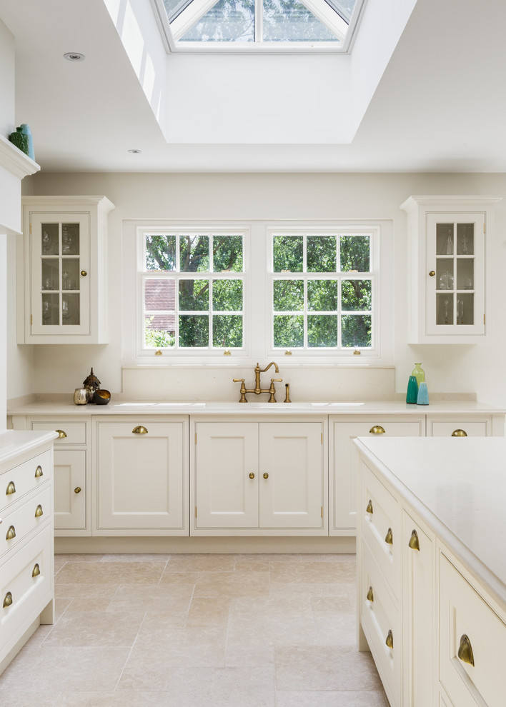 This is an example of a traditional kitchen in Cheshire with a farmhouse sink, beaded inset cabinets, white cabinets, quartzite benchtops, white splashback, glass sheet splashback, white appliances, limestone floors, with island, white floor and white benchtop.