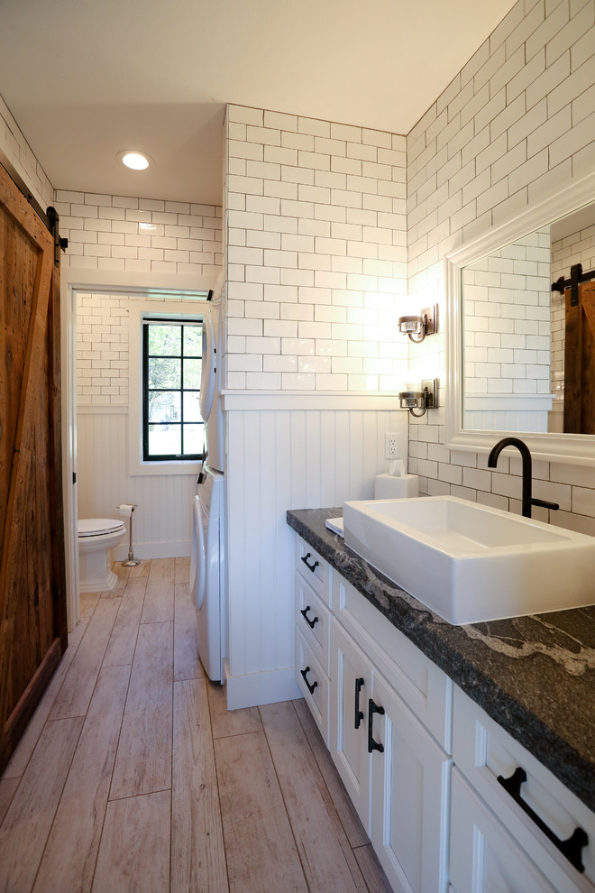 Photo of a mid-sized transitional master bathroom in Other with recessed-panel cabinets, white cabinets, an open shower, a two-piece toilet, white tile, subway tile, white walls, light hardwood floors, a vessel sink, soapstone benchtops and a laundry.