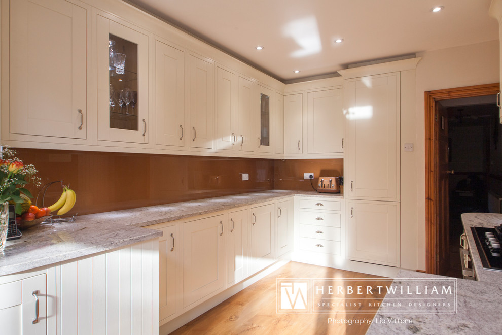 Large traditional l-shaped eat-in kitchen in Hampshire with shaker cabinets, white cabinets, glass benchtops, brown splashback, glass sheet splashback, stainless steel appliances, medium hardwood floors, a peninsula and brown floor.