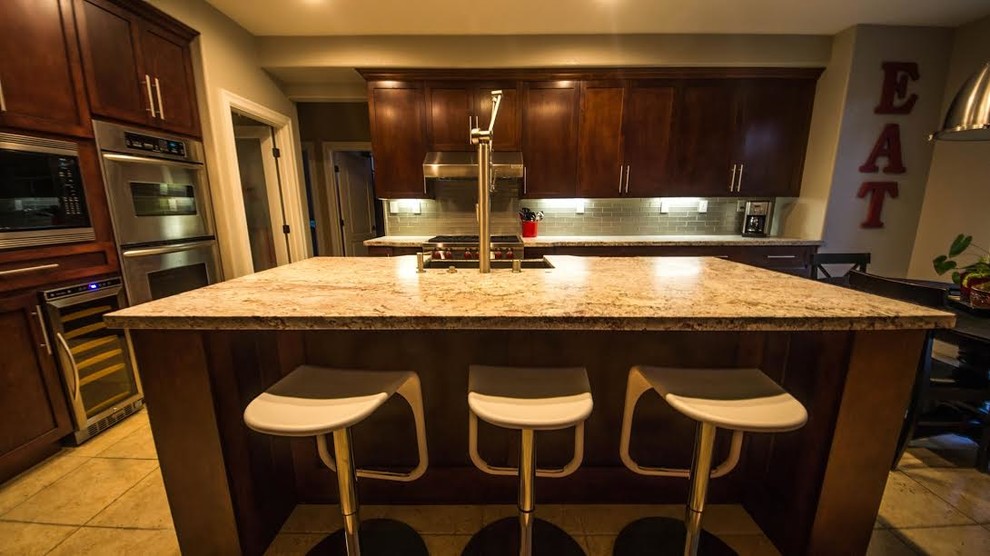 Eat-in kitchen - large transitional l-shaped ceramic tile eat-in kitchen idea with a farmhouse sink, shaker cabinets, brown cabinets, granite countertops, green backsplash, subway tile backsplash, stainless steel appliances and an island