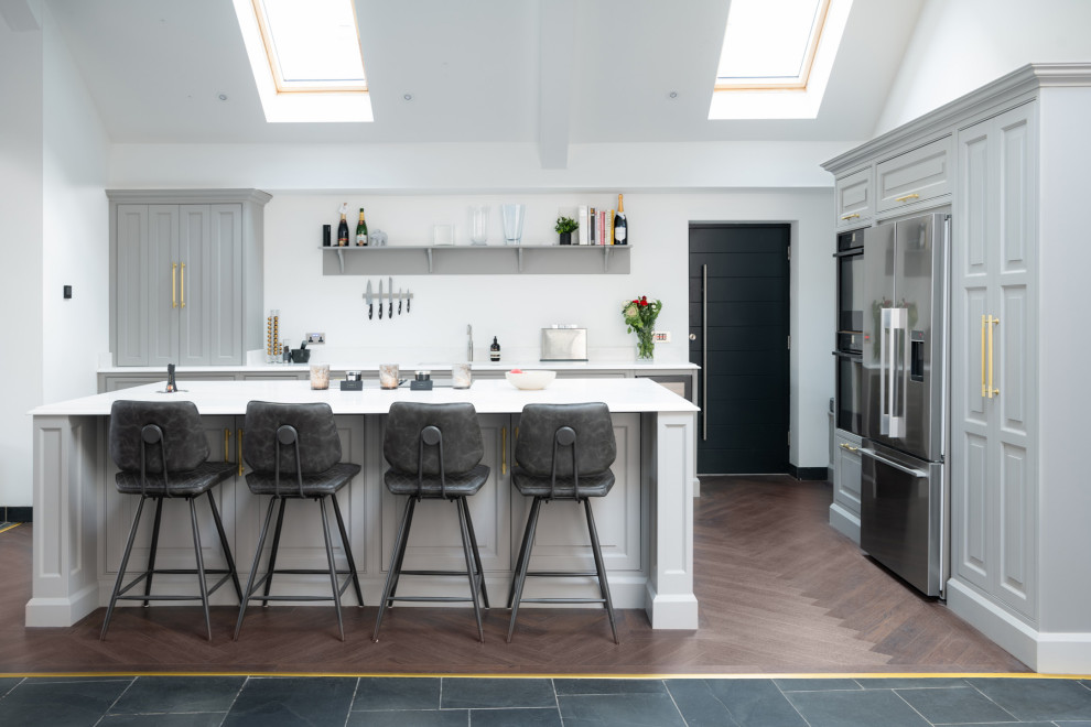 This is an example of an expansive classic grey and white galley open plan kitchen in Essex with an integrated sink, raised-panel cabinets, grey cabinets, quartz worktops, black appliances, laminate floors, an island, brown floors, white worktops, a vaulted ceiling and a feature wall.