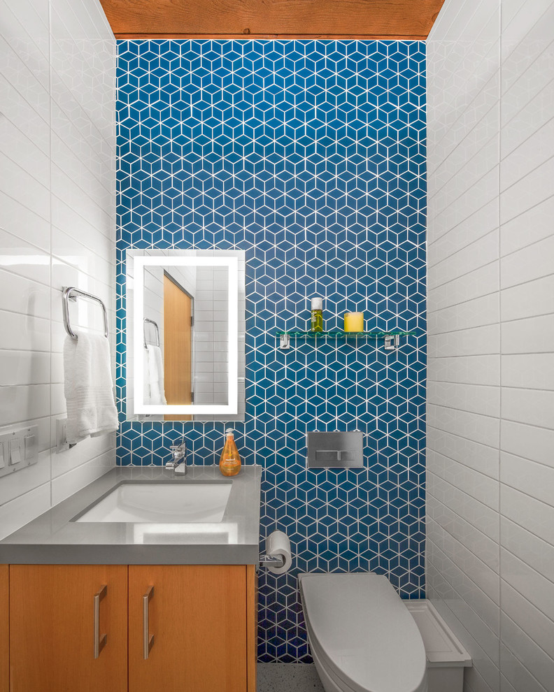 Inspiration for a midcentury powder room in Los Angeles with ceramic tile, flat-panel cabinets, medium wood cabinets, a wall-mount toilet, white tile, blue tile, blue walls, an undermount sink and grey benchtops.