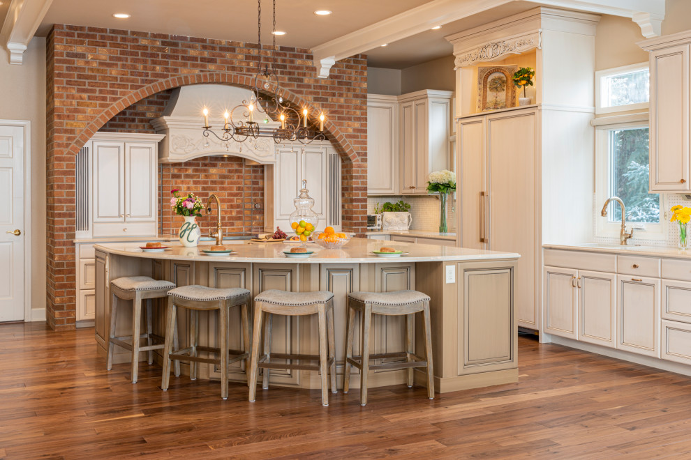 Inspiration for a l-shaped kitchen in Denver with quartz benchtops, brick splashback, medium hardwood floors, with island, recessed-panel cabinets, beige cabinets, panelled appliances, brown floor and beige benchtop.