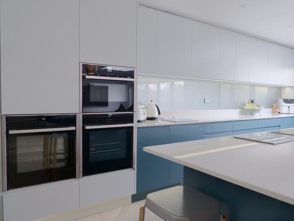 Photo of a large contemporary l-shaped open plan kitchen in London with a built-in sink, flat-panel cabinets, green cabinets, quartz worktops, grey splashback, glass sheet splashback, stainless steel appliances, light hardwood flooring, an island, brown floors and white worktops.
